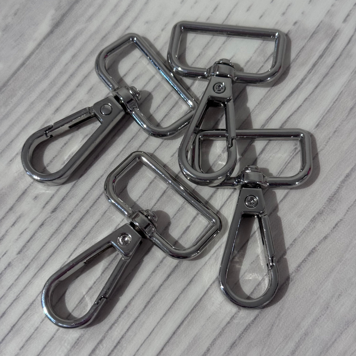 Four shiny silver metal swivel snap hooks with rectangular loops, arranged on a light gray wood-textured surface.