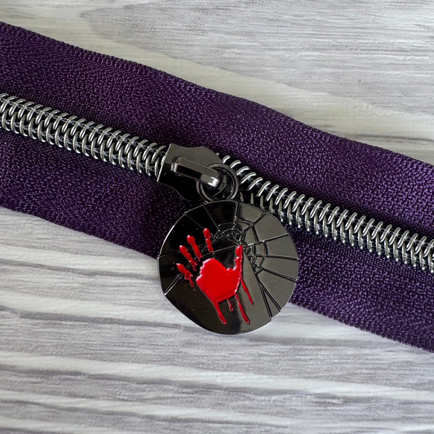 Close-up of a purple zipper with a black zipper pull featuring a red handprint design on a gray and white striped background.